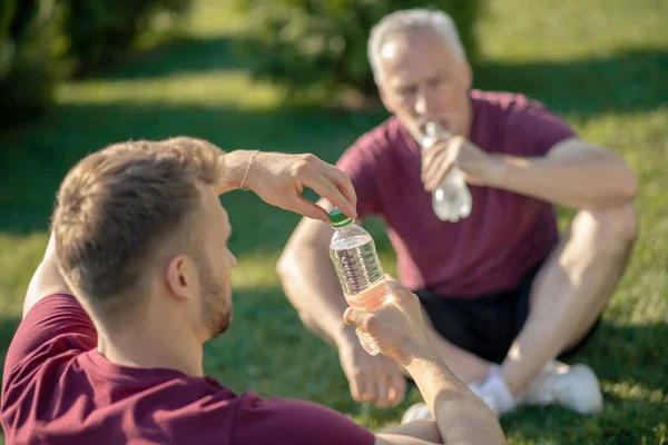 Bearded male opening bottle of water, grey-haired man drinking water, both sitting on grass — Stock Photo, Image