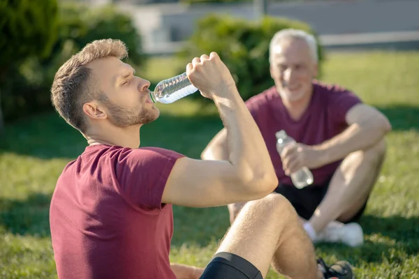 Бородатый мужчина пьет воду, седой мужчина держит бутылку, оба сидят на траве — стоковое фото
