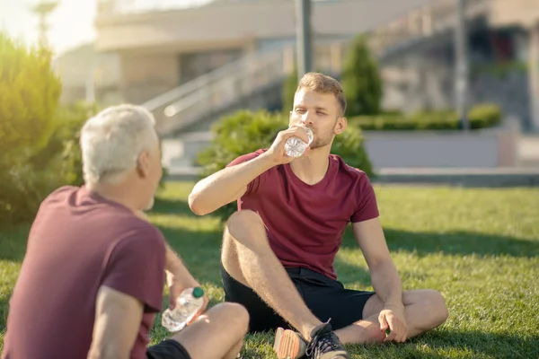 Молодой мужчина пьет воду, взрослый мужчина держит бутылку, оба сидят на траве — стоковое фото