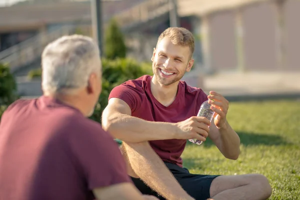 Бородатый мужчина закрывает бутылку, улыбается, седой мужчина сидит на траве напротив него — стоковое фото