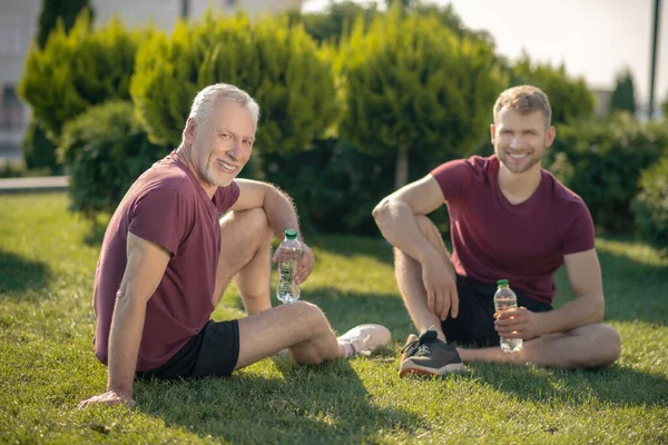 Молодой мужчина и зрелый мужчина сидит на траве, пьет воду, улыбается — стоковое фото