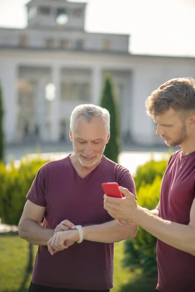Ώριμος άντρας ελέγχει SmartWatch με νέους άνδρες εκμετάλλευση smartphone — Φωτογραφία Αρχείου