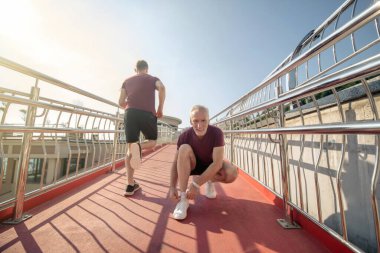 Gri saçlı erkek, spor ayakkabılarını bağlıyor, genç erkek arkasından koşuyor.