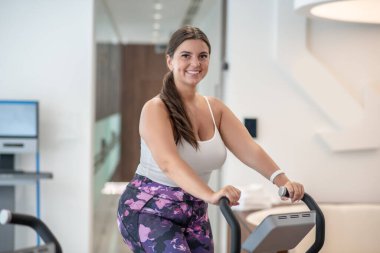 Spor salonunda egzersiz bisikleti kullanan gülümseyen bir kadın.