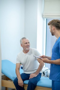 Genç sakallı doktor yetişkin erkek hastaya klip gösteriyor.
