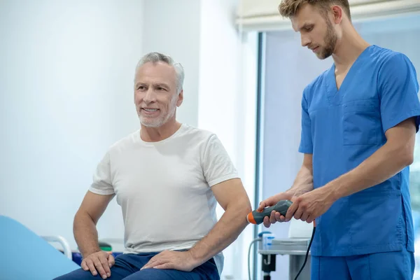 Paziente maschio dai capelli grigi sottoposto a trattamento ecografico al gomito, seduto sul pullman — Foto Stock