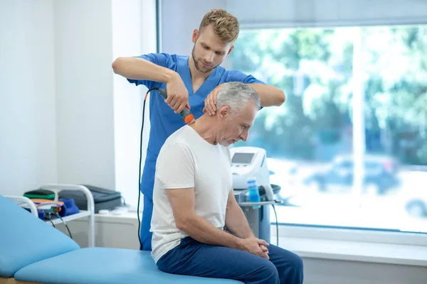Paciente masculino de pelo gris sentado en el sofá, recibiendo tratamiento de ultrasonido en el cuello —  Fotos de Stock