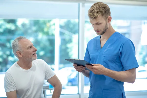 Bearded doctor standing with clip folder, talking to mature male patient — Stock Photo, Image