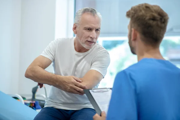 Paciente varón maduro mostrando codo al médico de cabello castaño —  Fotos de Stock