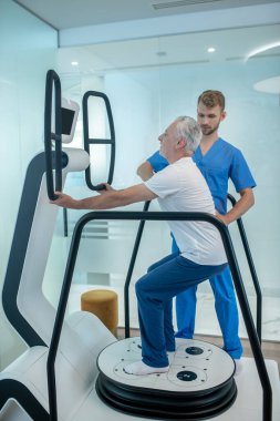 Gri saçlı adam egzersiz makinesi ve doktorun yanında çömeliyor.
