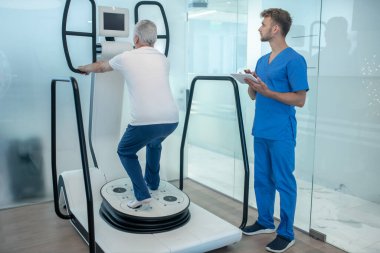 Simülatördeki yaşlı adam ve tabletli rehabilitasyon uzmanı