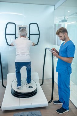 Simülatör yanında gri saçlı adam ve tabletli eğitmen.