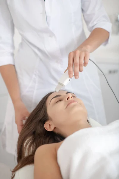 Ciemnowłosa kobieta leżąca na kanapie podczas laserowego leczenia skóry w salonie — Zdjęcie stockowe