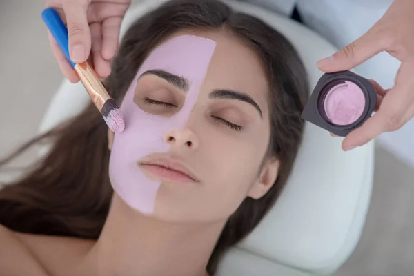 Kosmetologis menerapkan masker wajah merah muda pada wajah wanita — Stok Foto