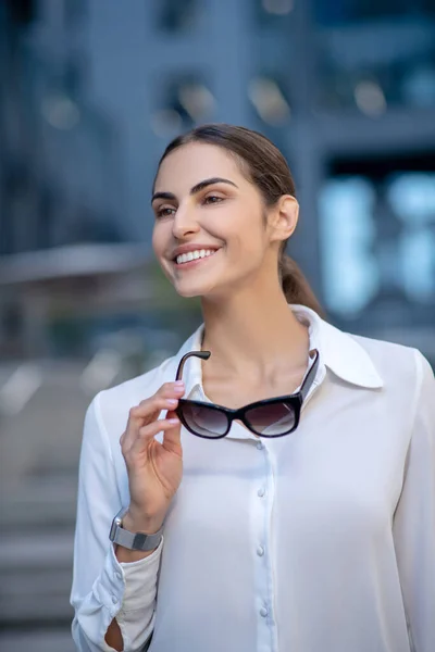 Жінка в білій сорочці тримає сонцезахисні окуляри і посміхається — стокове фото