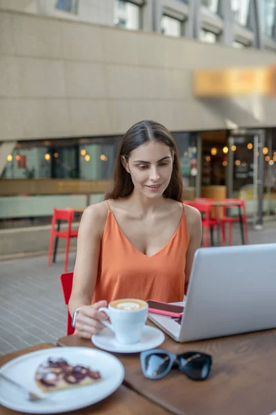 Tmavovlasá hezká žena si dává kávu a dívá se na něco na notebooku — Stock fotografie