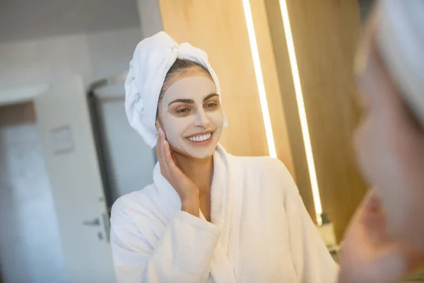 Ung kvinna med ansiktsmask tittar i spegeln och ler — Stockfoto