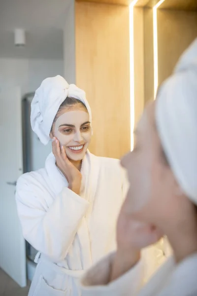 Wanita muda dengan masker wajah melihat di cermin dan tampak bahagia — Stok Foto