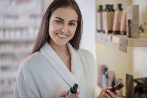 Mujer en bata blanca sosteniendo productos de belleza y sonriendo — Foto de Stock