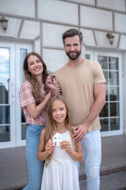 Evin yanında duran ve gülümseyen genç bir aile.