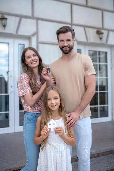 Evin yanında duran ve gülümseyen genç bir aile.