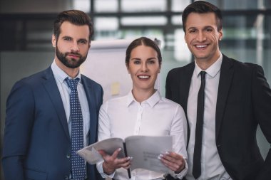 Kağıtları tutan kahverengi saçlı kadın, erkek iş arkadaşlarının arasında duruyor.