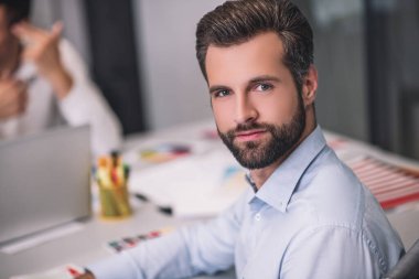 Sakallı erkek masada oturuyor, kameraya bakıyor.