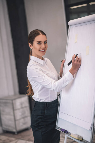 Smiling brown-haired female in white shirt writing on flipchart