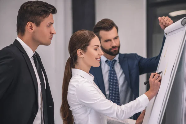 Bruinharige vrouw schrijft op flipchart, mannelijke collega 's bestuderen zorgvuldig notities — Stockfoto