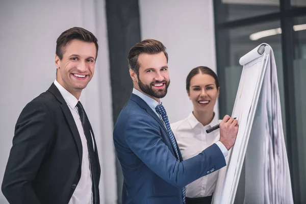 Skäggigt manligt skrivande på blädderblock, hans kvinnliga och manliga medarbetare står nära — Stockfoto