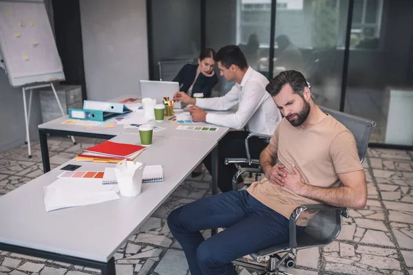 Varón barbudo cansado sentado en la reunión, tocando su estómago — Foto de Stock