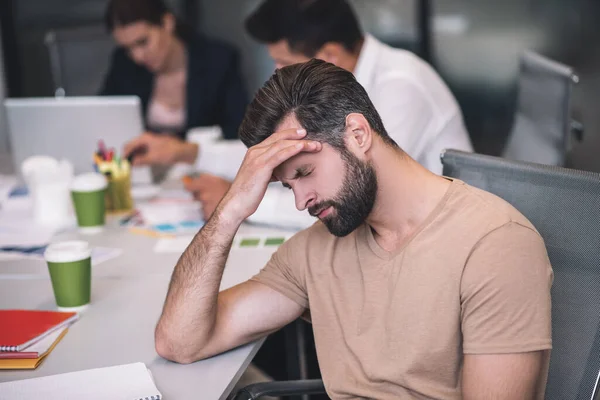 Bearded male sitting at meeting, touching his forehead