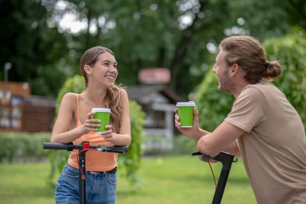 Два человека со скутерами разговаривают в парке и пьют кофе вместе — стоковое фото