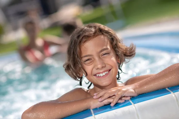 Boy holding swimming pool border, his parents swimming behind — Stock Photo, Image