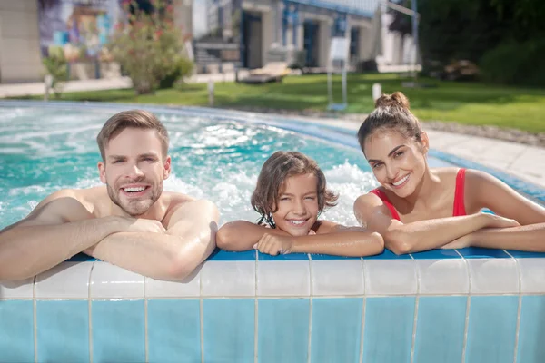 Family of three holding swimming pool border, smiling — Stock Photo, Image