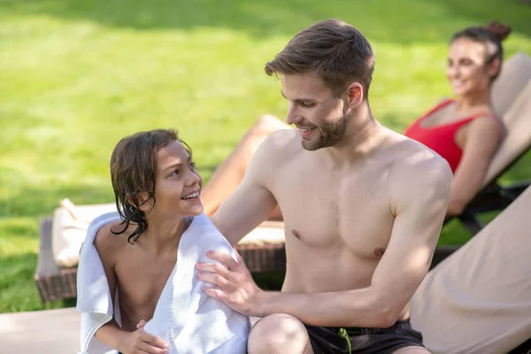 Father wrapping his son in towel, his wife resting on sun lounger — Stock Photo, Image