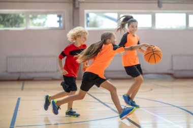 Parlak spor giyimli çocuklar birlikte basketbol oynuyorlar ve rekabetçi hissediyorlar.
