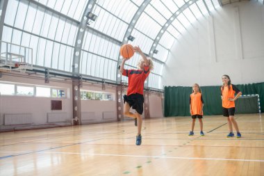 Kırmızı tişörtlü sarışın çocuk elinde topla zıplıyor.