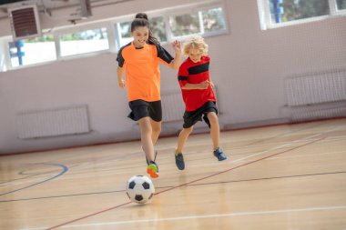 Spor giyimli üç çocuk spor salonunda topun peşinden koşuyor.
