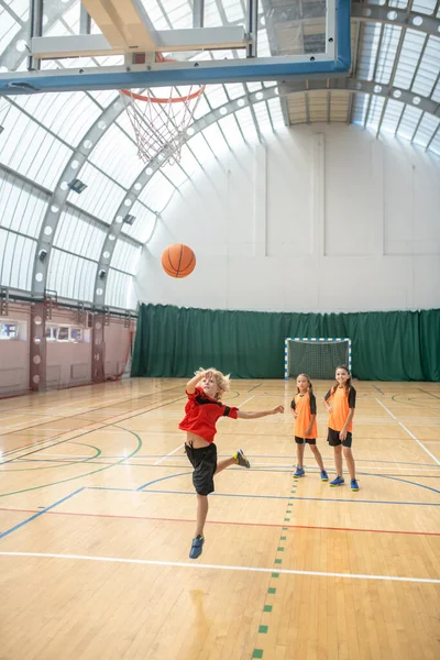 Kırmızı tişörtlü sarışın çocuk topu sepete atıyor.