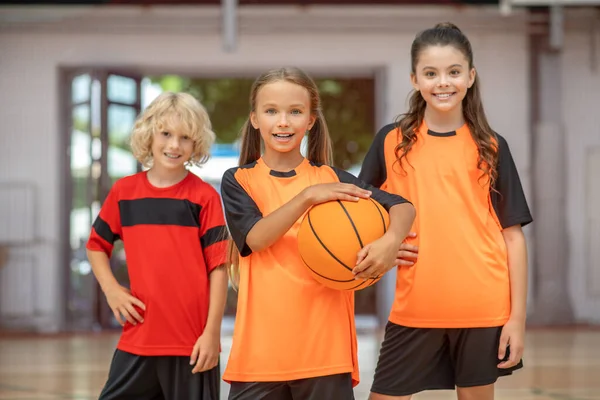 Spor kıyafetli çocuklar elinde bir topla mutlu bir şekilde gülümsüyor.