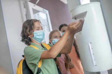 Koruyucu maskeli çocuk eline dezenfektan alıyor.