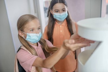 Sarı saçlı, koruyucu maskeli bir kız eline dezenfektan alıyor.