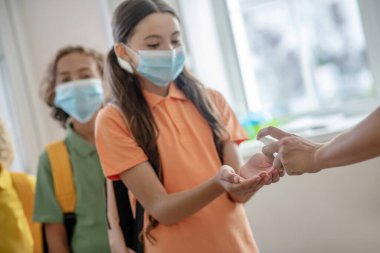 Siyah saçlı, koruyucu maskeli bir kız eline dezenfektan alıyor.