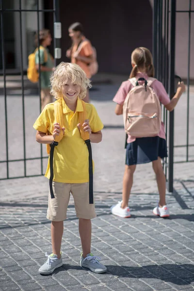 Lindo colegial con una mochila sonriendo positivamente mientras está de pie cerca de la entrada de la escuela — Foto de Stock