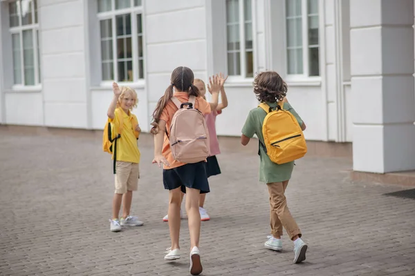 Los escolares con máscaras protectoras en el patio de la escuela saludándose saludándose saludando con las manos —  Fotos de Stock