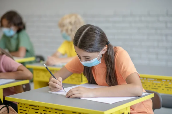 Fille en masque écrit dans un cahier et regardant impliqué — Photo