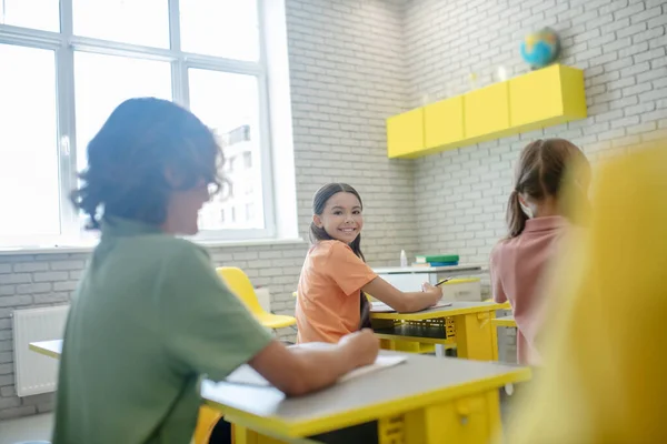 Menina de cabelos escuros sentada na mesa na sala de aula e sorrindo para sua colega de classe — Fotografia de Stock