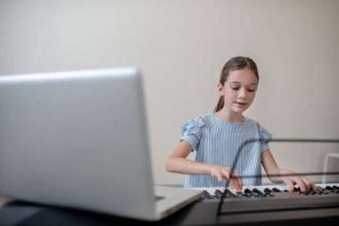 Uzun saçlı, mavi elbiseli kız sentezleyicinin önünde oturmuş konsantre olmuş görünüyor.