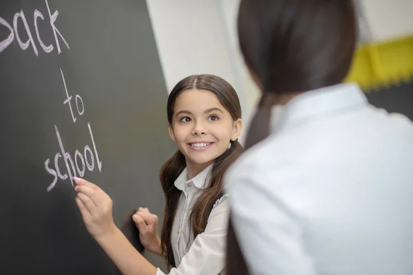 Улыбающаяся милая девушка пишет на доске и смотрит на своего учителя — стоковое фото